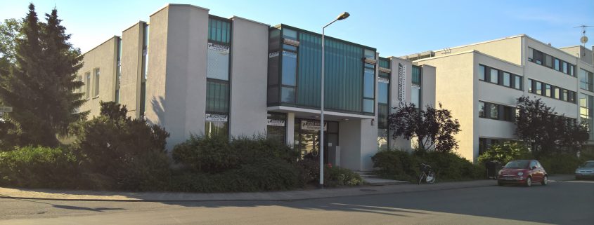 La photo montre un immeuble moderne abritant des bureaux et des salles de formation, situé à l'angle d'une rue, prise lorsque le soleil était bas. La façade est claire et sobre, percée de grandes baies vitrées encadrées de vert qui confèrent au bâtiment un caractère à la fois sobre et marquant. Au-dessus de l'entrée est accrochée une enseigne portant l'inscription « STUDIO MONDIALE Sprachenschule » (école de langues), d'autres enseignes avec le nom sont apposées sur les fenêtres. Devant le bâtiment se trouve une rue calme, au bord de laquelle sont garées une petite voiture rouge et un autre véhicule. À droite de l'entrée, un vélo est appuyé contre le trottoir. Devant la maison poussent des buissons et deux petits arbres au feuillage sombre, tandis qu'à gauche, un grand sapin s'élève vers le ciel.